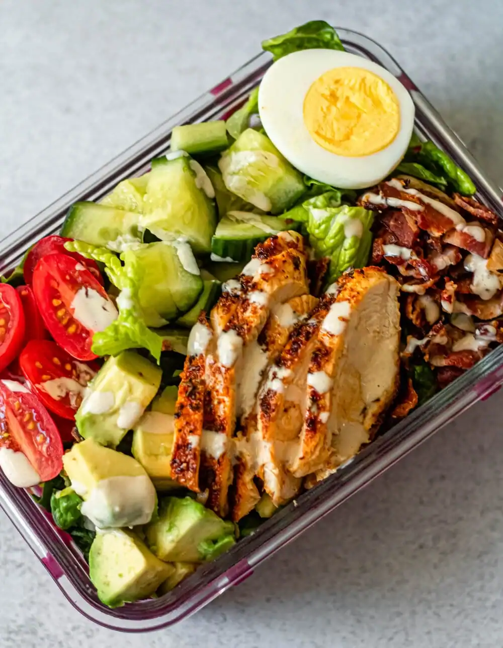 Meal prepped Cobb salad with cucumbers, cherry tomatoes, hard-boiled egg, chicken and bacon in a glass container