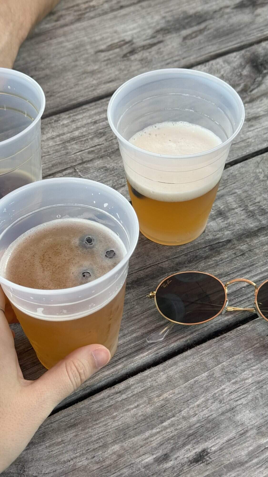Three beers in plastic cups on an outdoor table, next to some Ray Ban sunglasses.