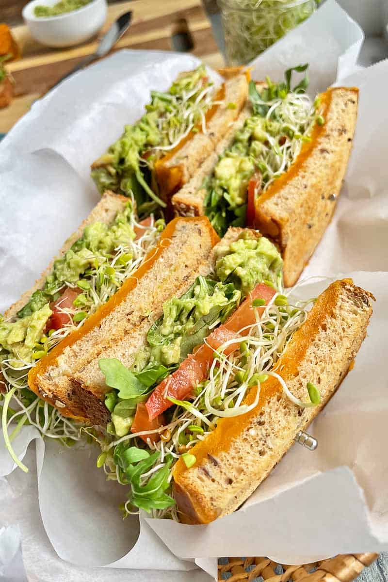 Fresh veggie and avocado toasted sandwich with sprouts, tomatoes, and melted cheese on whole grain bread, served with a side of shredded lettuce. Perfect for a healthy and delicious lunch.