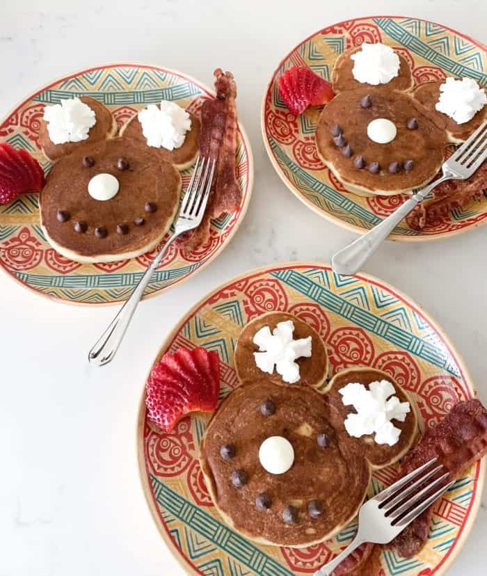 Cute reindeer pancake breakfast with chocolate chips, whipped cream, strawberries, and bacon on colorful decorative plates, perfect for a festive holiday morning.