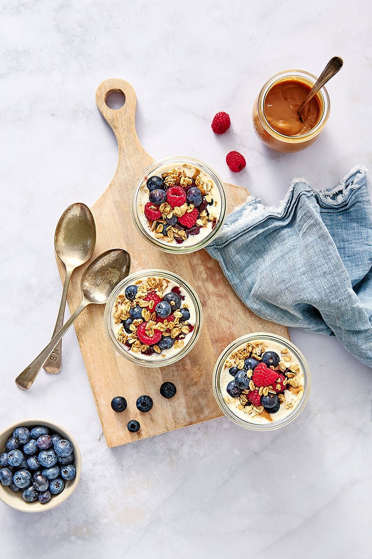 Overhead shot of creamy overnight oats with no chia seeds in glass jars, topped with berries and sitting on a wooden board next to spoons and peanut butter.