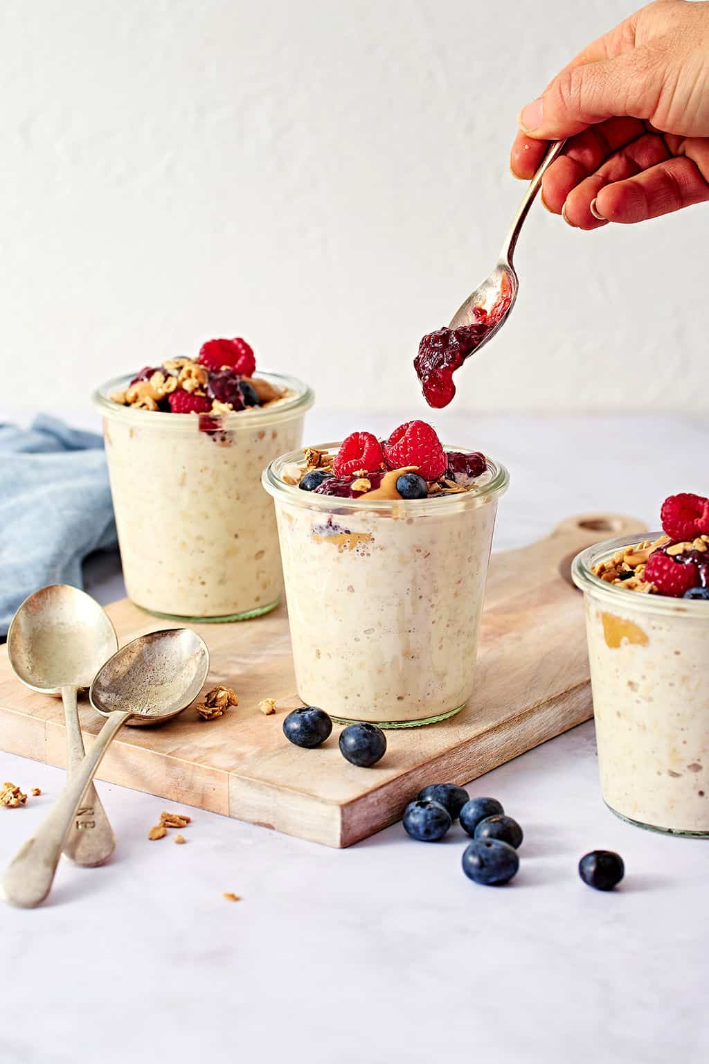 Creamy overnight oats in glass jars, being topped with fruit jam, perfect for breakfast or snacks.