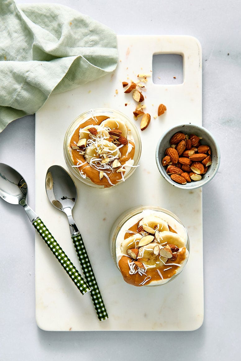 Overhead view of overnight oats oats with coconut milk topped with sliced bananas, shredded coconut, and almonds on a white marble cutting board with spoons, nuts, and a light green cloth for a delicious breakfast presentation.