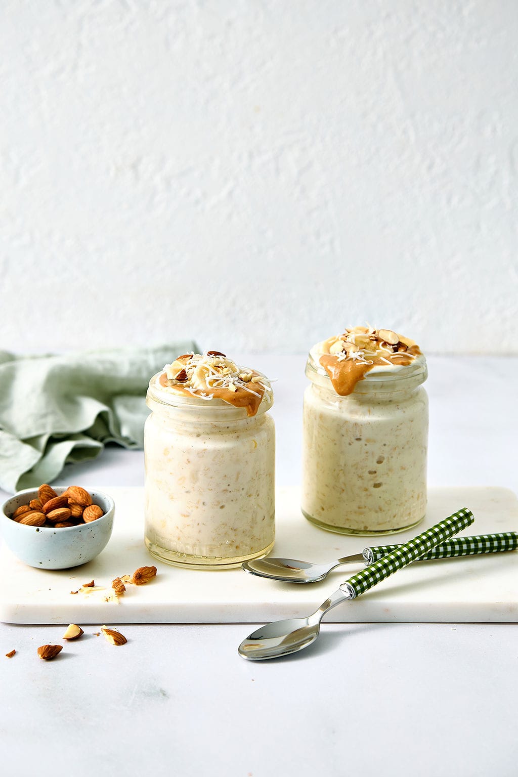 Creamy overnight oats with coconut milk topped with peanut butter and chopped almonds, served in glass jars with fresh almonds on a white marble slab. Perfect for healthy meal prep or breakfast ideas.