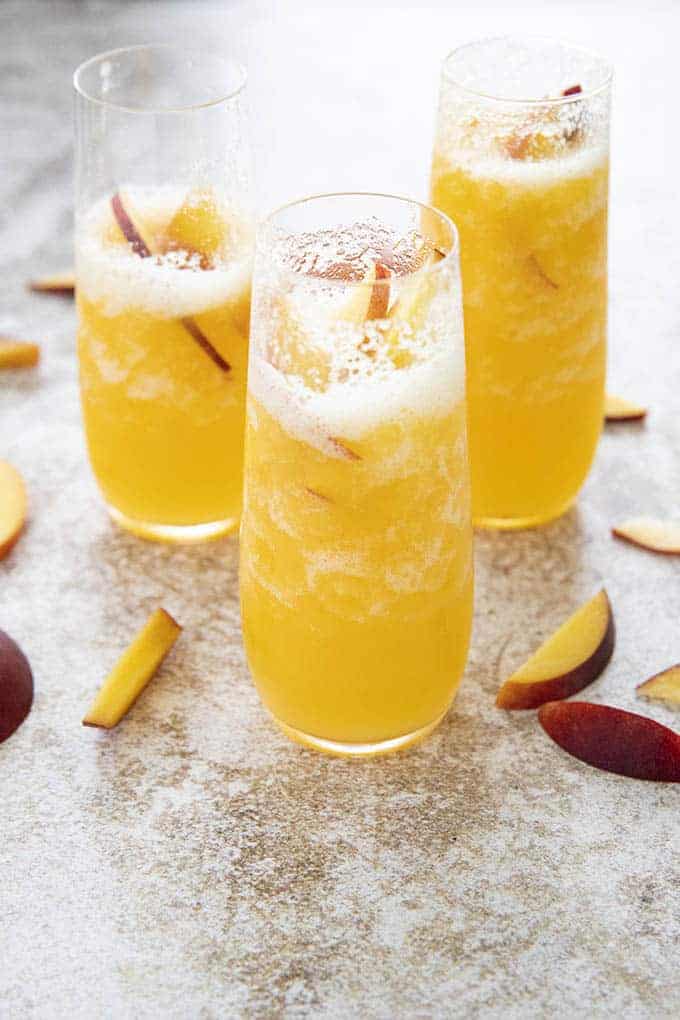 Three bellinis in champagne flutes, surrounded by peach slices.
