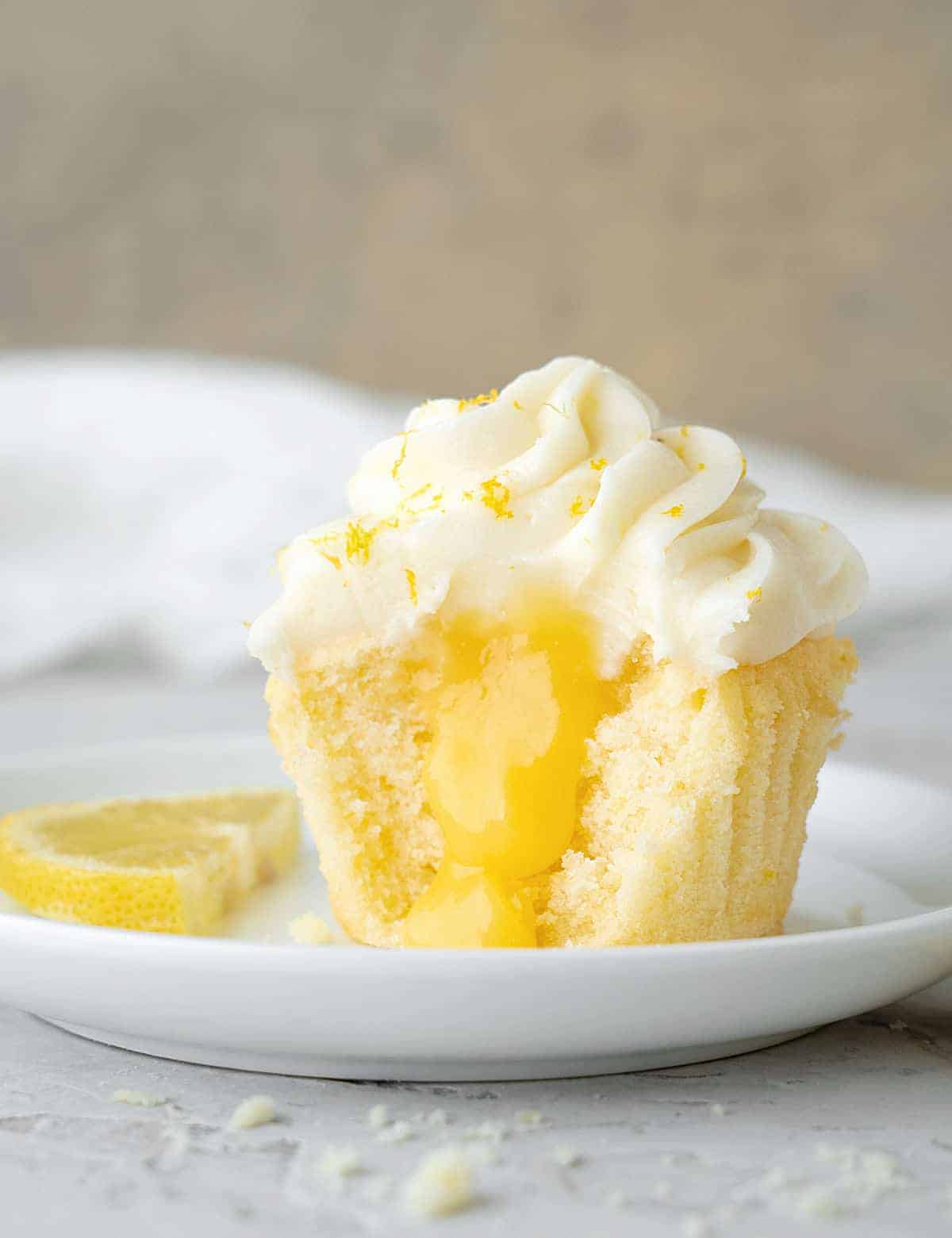 A dainty yellow cupcake filled with bright yellow lemon curd and topped with piped white icing and lemon zest.