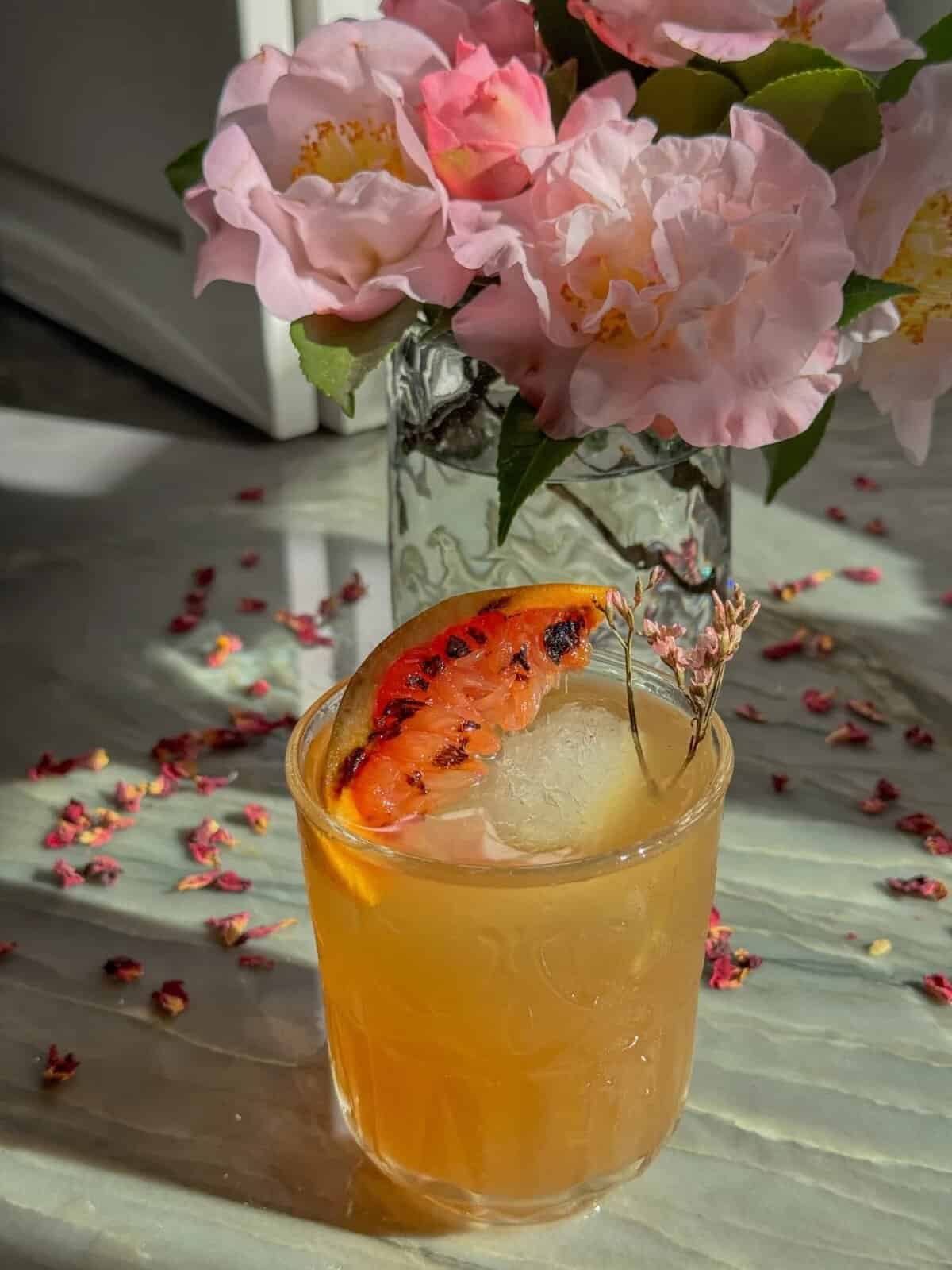 A rich, orange cocktail served in a rocks glass with a large ice cube, garnished with grilled grapefruit slice and small edible flower stem, presented in front of a vase of pink flowers.