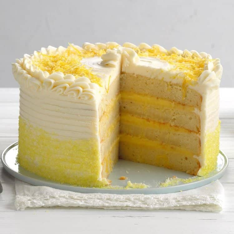 A tall lemon layer cake with a large slice taken to showcase the interior, topped with white icing, lemon zest and a piped icing rim.