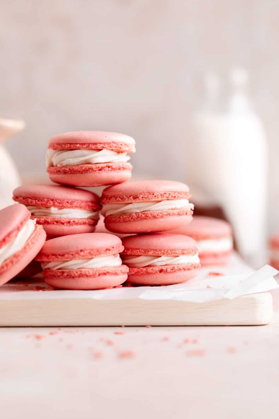 Pink macarons with white filling stacked on a white wooden tray, with a blurred background featuring a milk bottle.