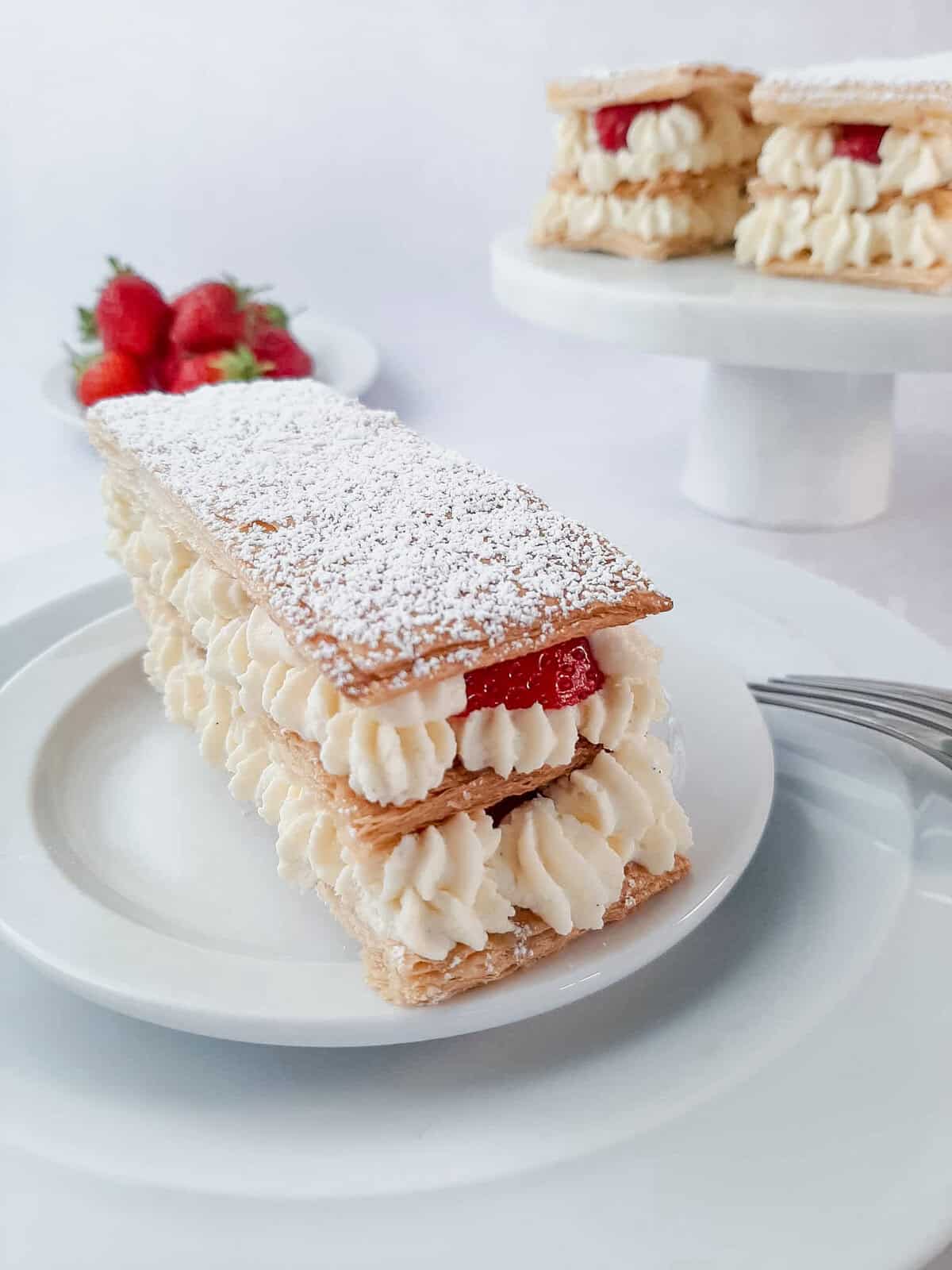 Layered Mille Feuille pastry with cream and fresh strawberries, dusted with powdered sugar, served on a white plate.