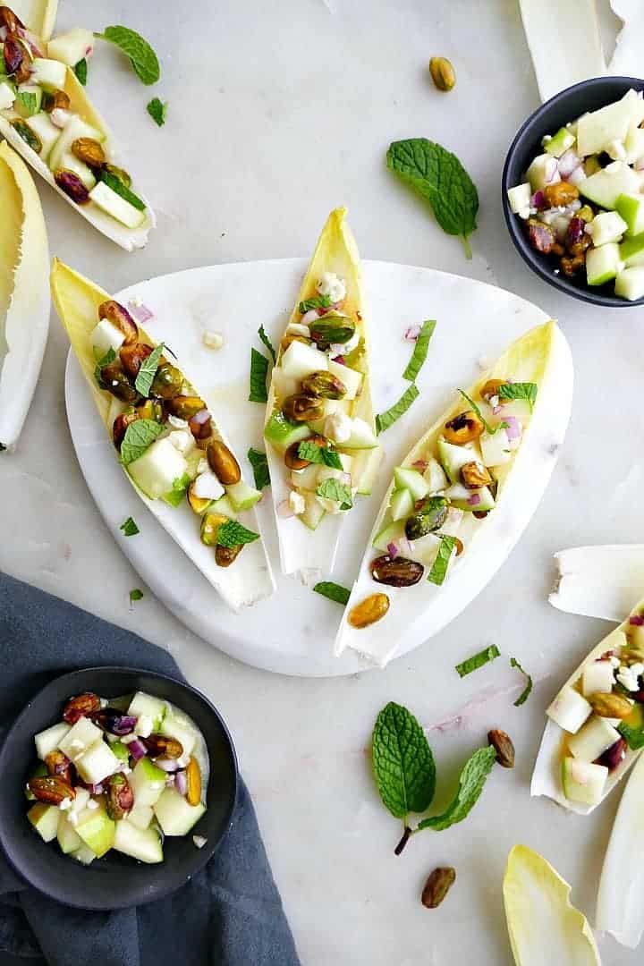 Light green endive leaves filled with diced apples, pistachios and crumbled blue cheese, on a triangular, white marble serving plate.