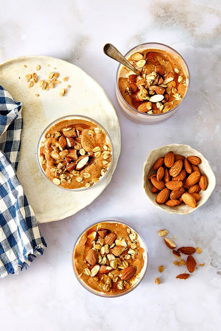 Creamy almond butter overnight oats topped with crunchy mixed nuts in glass cups, served on a marble table next to a small bowl of almonds and a blue checkered cloth.