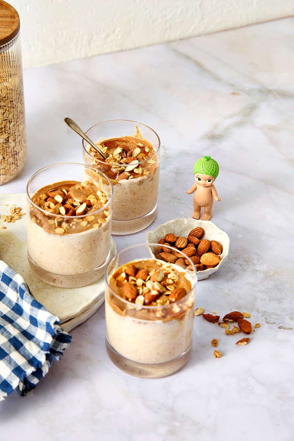 Creamy almond butter overnight oats topped with chopped nuts in glass cups, being admired by a small Sonny Angel doll figure standing next to a bowl of almonds on a marble table.