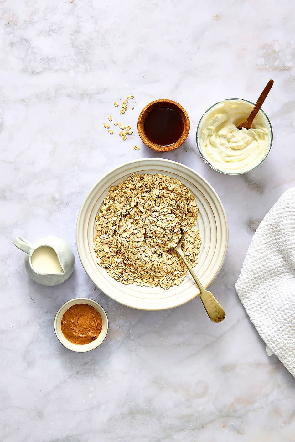 Overnight oats ingredients including yogurt, oats, honey, and almond butter on a light marble surface.