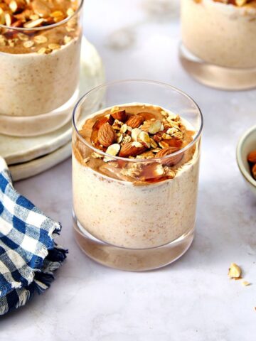 Creamy almond butter overnight oats topped with chopped nuts in a clear glass, served with a bowl of almonds and a blue checkered cloth on a marble table.