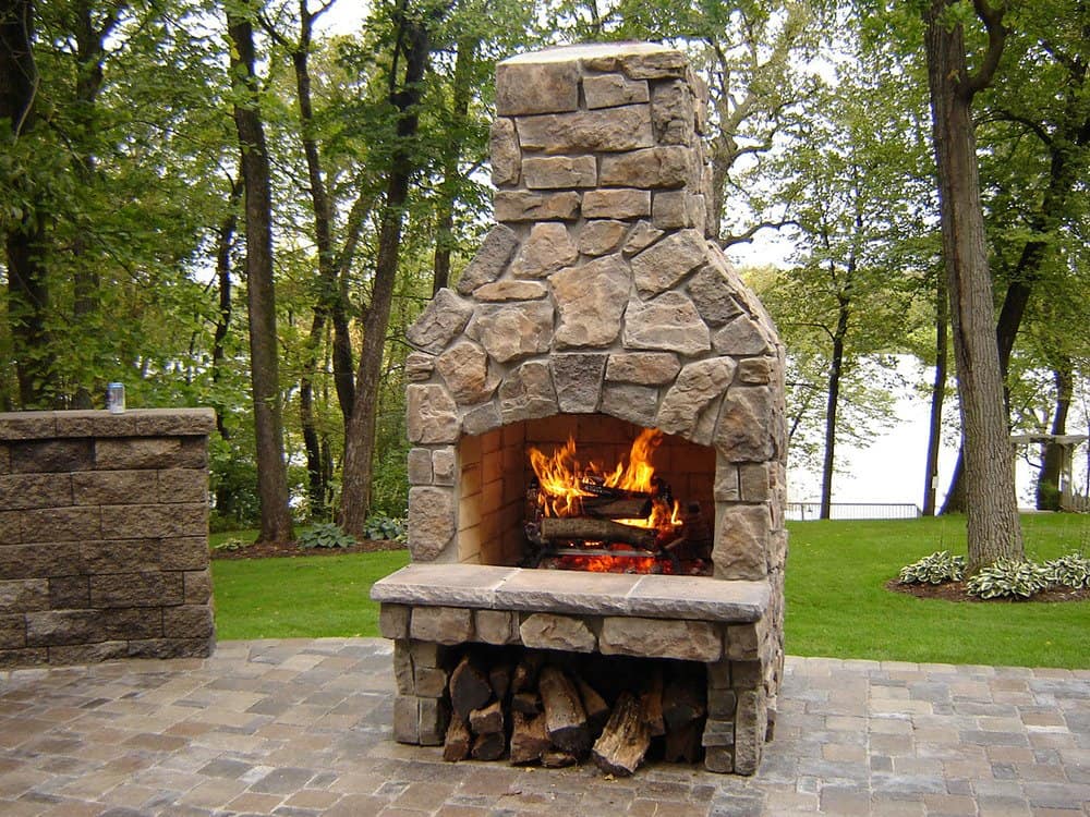 Crushed stone outdoor fireplace with fire in a landscaped backyard.
