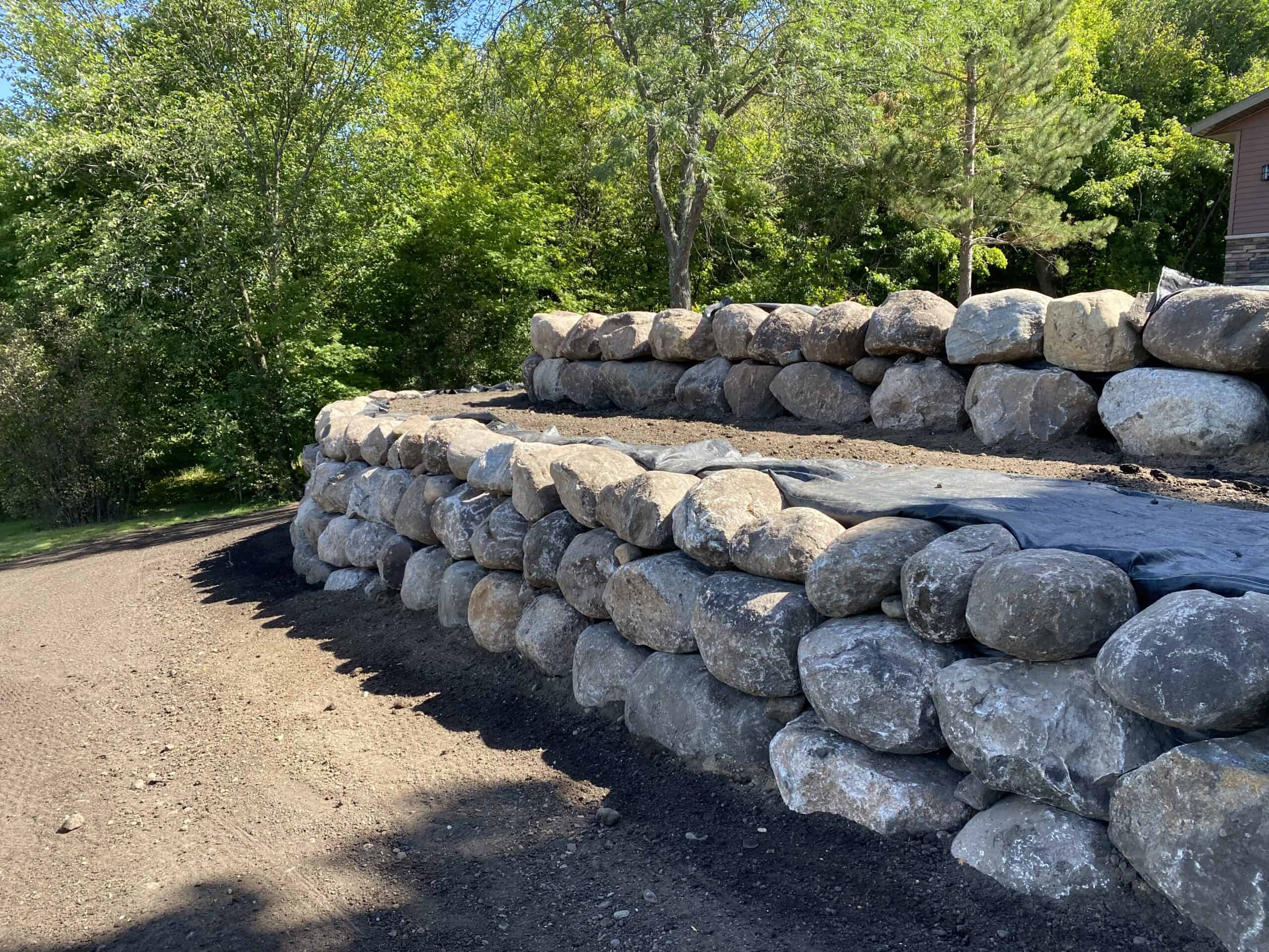 Rock retaining wall construction with large stones, landscape erosion control, and landscaping services in progress.