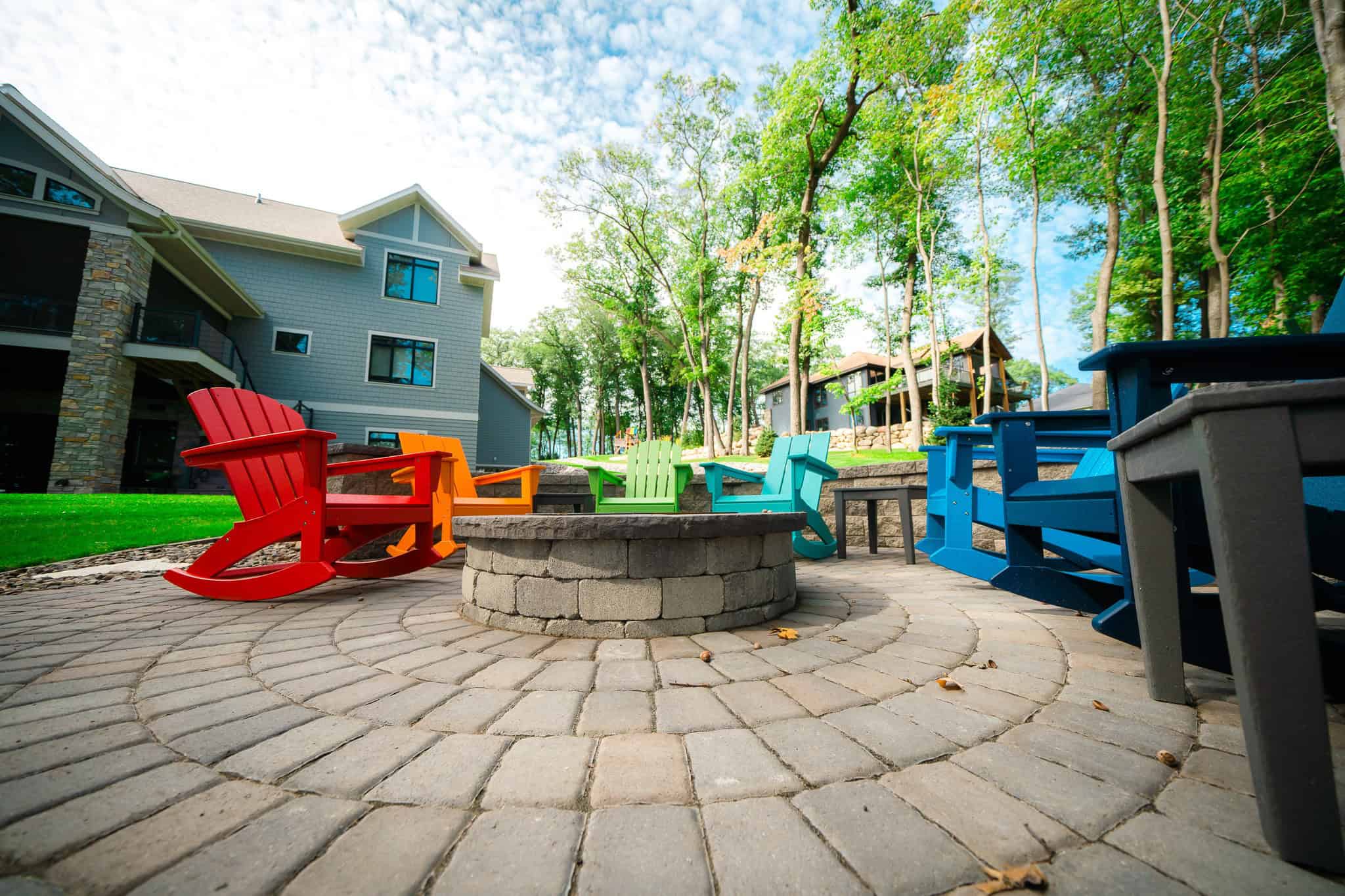 Colorful outdoor Adirondack chairs around a fire pit on a landscaped patio with lush green trees.