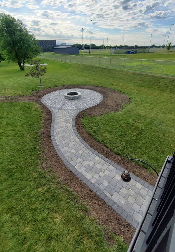 Paved backyard pathway with fire pit, landscaped grass, and outdoor lighting for garden enhancement.