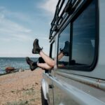Beachside vehicle with legs hanging out of window, sunny day, ocean view.