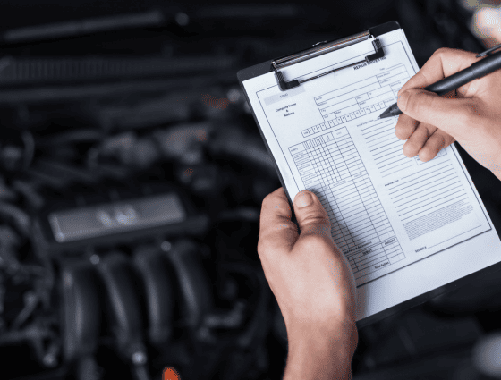 Technician inspecting vehicle engine with clipboard at auto repair shop.