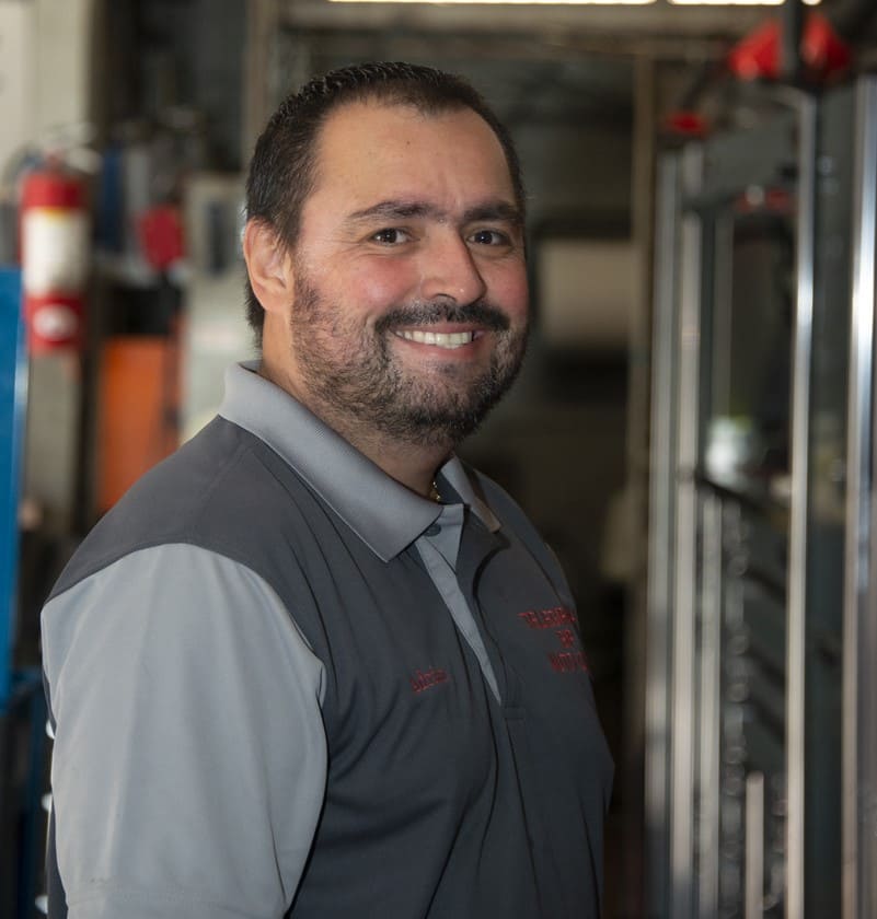 Auto repair technician smiling in workshop with tools and car parts in background.