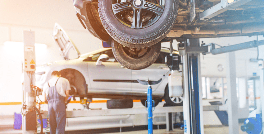 Auto repair shop with a car lifted on a hydraulic lift, mechanic working on the vehicle's underside.