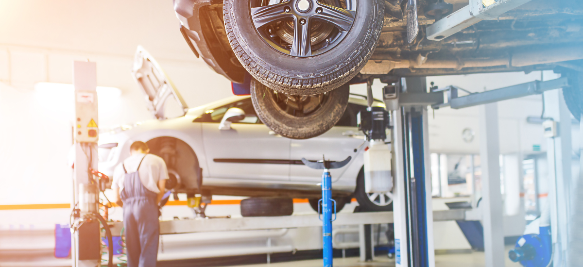 Auto repair shop with a car lifted on a hydraulic lift, mechanic working on the vehicle's underside.