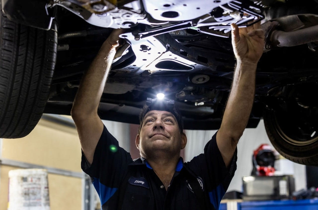 Auto mechanic inspecting vehicle undercarriage in repair shop.