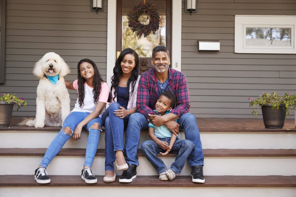 An image that shows Family With Children And Pet Dog Sit On Steps Of Home