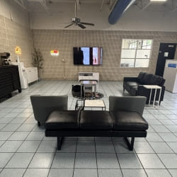 Modern auto shop lounge with black leather sofas, TV, and tiled flooring for customer comfort.
