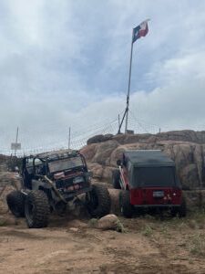 Off-road vehicle navigating rocky terrain with Texas flag in background.