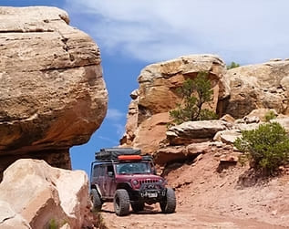 Red Jeep navigating rugged desert terrain with large rock formations in the background.