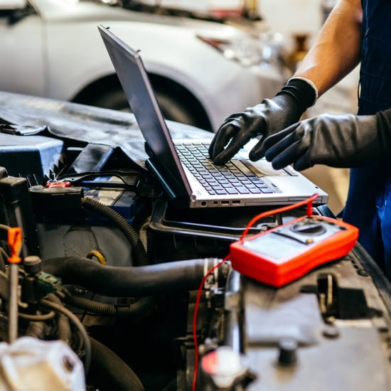 Automotive technician using a laptop for engine diagnostics under the hood of a car.