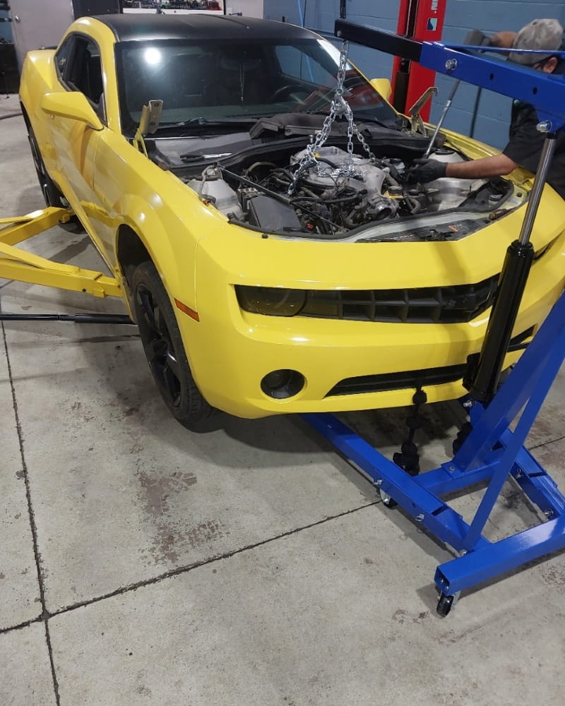 Engine repair on a yellow Chevrolet Camaro with engine hoist and mechanic.