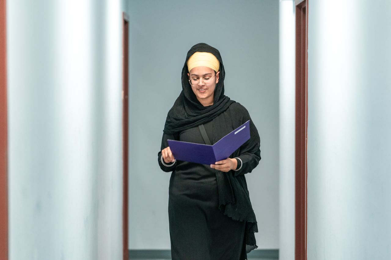 Smiling businesswoman wearing hijab and glasses walks confidently through a corridor holding a folder.