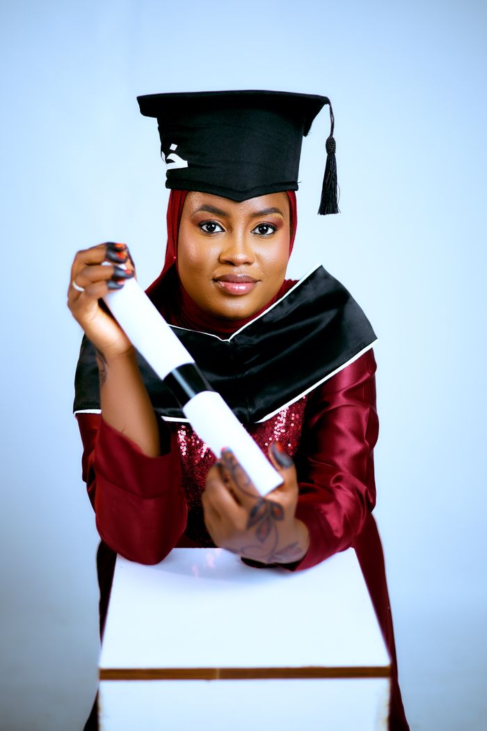 Young graduate wearing a cap and gown, proudly holding a diploma against a light background.
