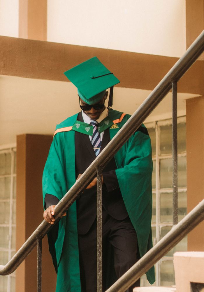 A young graduate in green academic attire descends a staircase outdoors.