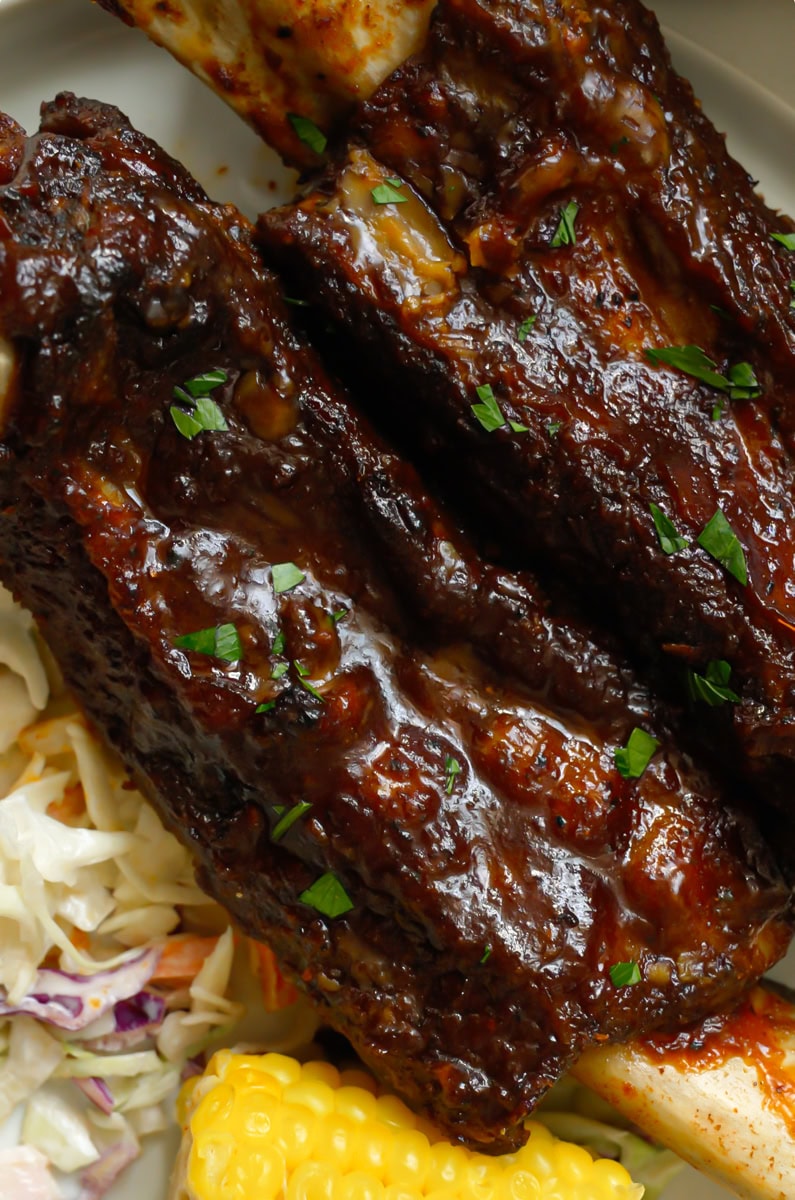 Juicy tender oven baked beef ribs with bbq sauce on a plate with southern coleslaw and buttered corn.