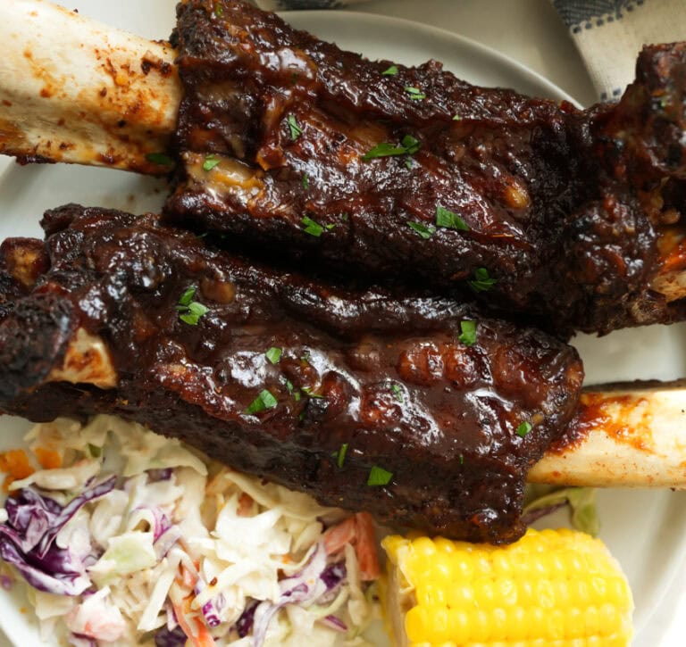 A picture of oven baked beef ribs with bbq sauce on a plate with southern coleslaw and buttered corn.