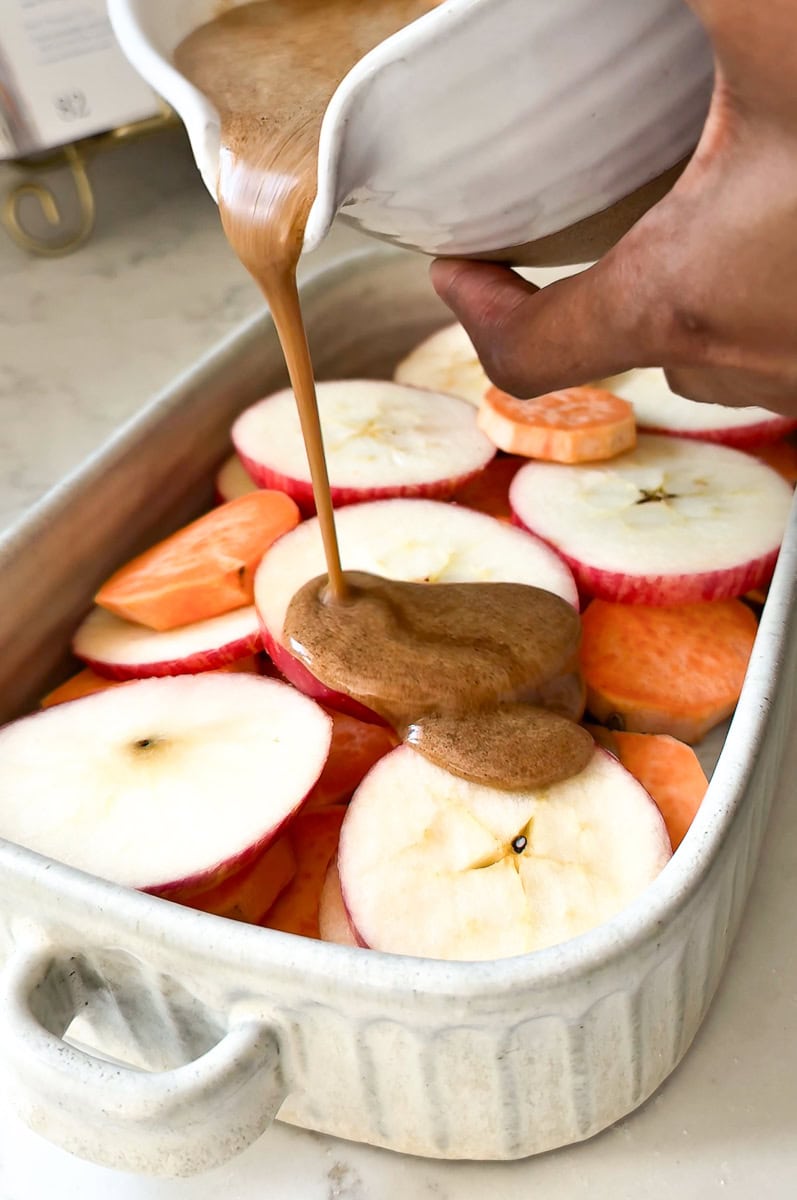 Raw apple and sweet potato slices layered in a baking dish with melted brown sugar mixture being poured on top.