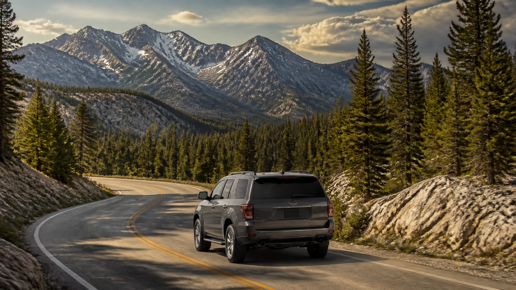 SUV driving on a mountain road surrounded by pine trees and mountains.
