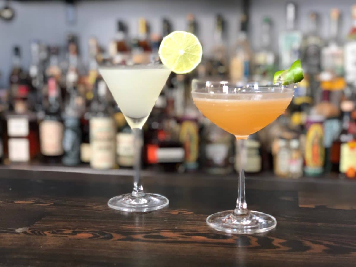 Two cocktails on a wooden bar, one light-colored with a lime slice, the other orange with a green garnish
