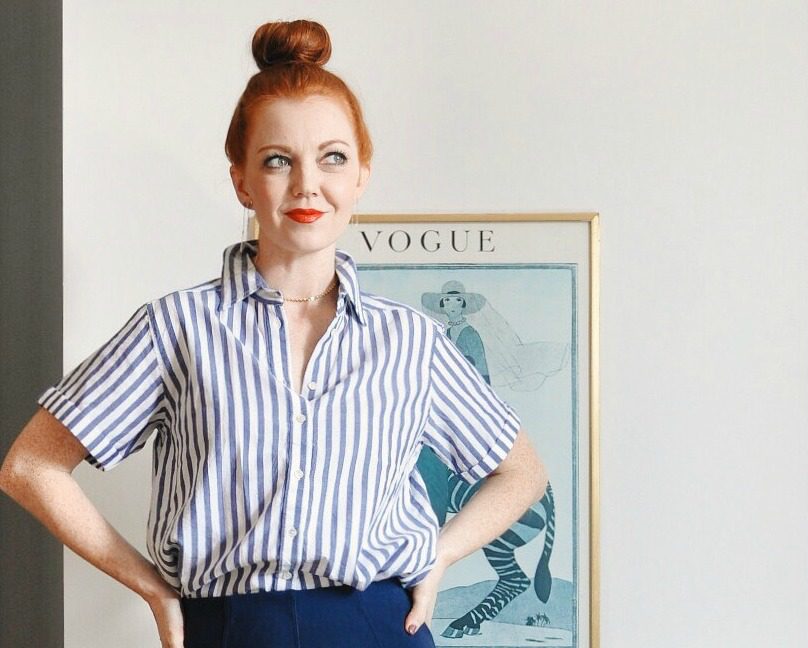 A woman with red hair in a bun, wearing a blue and white striped shirt and dark skirt, stands looking right