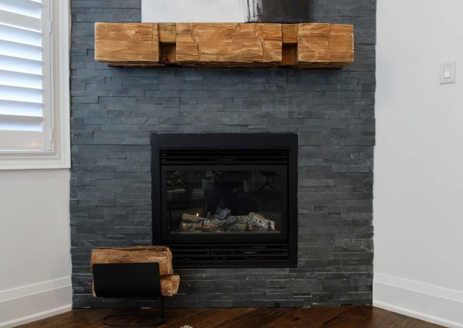 Rustic fireplace with dark stacked stone, chunky wood mantel, and firewood holder on hardwood flooring