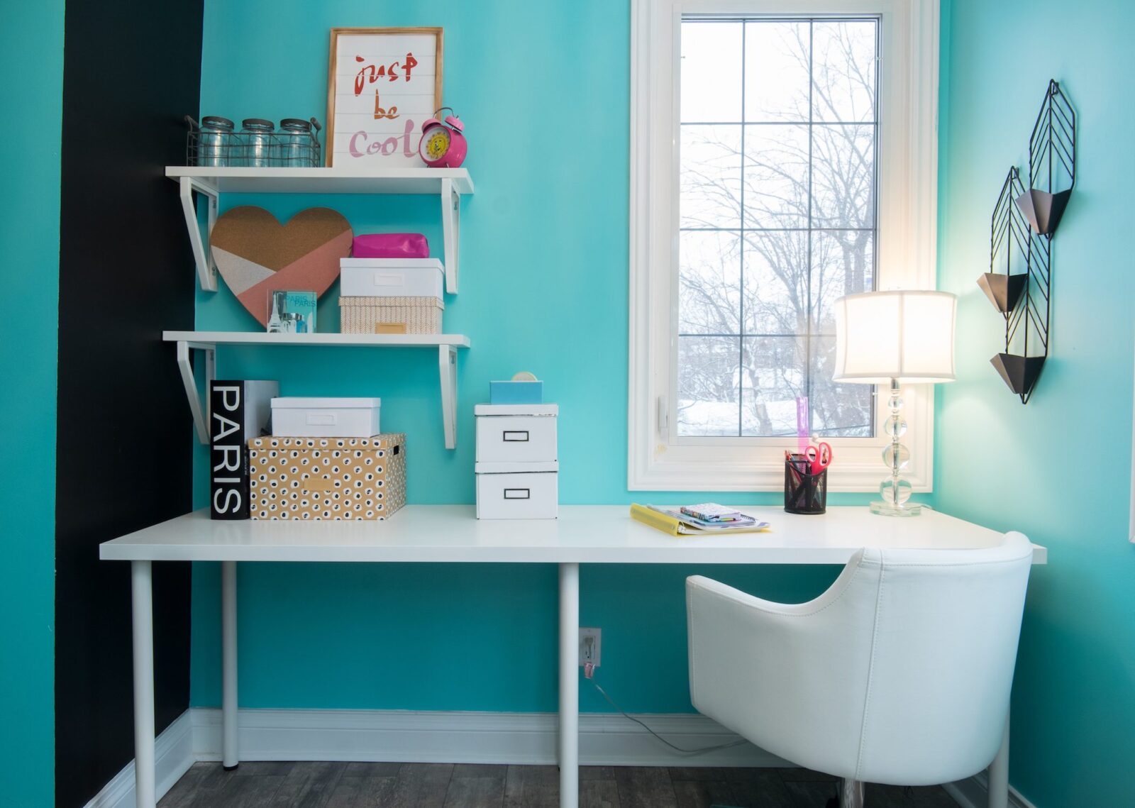 Bright turquoise home office with white desk, floating shelves, storage boxes, and a window-side lamp