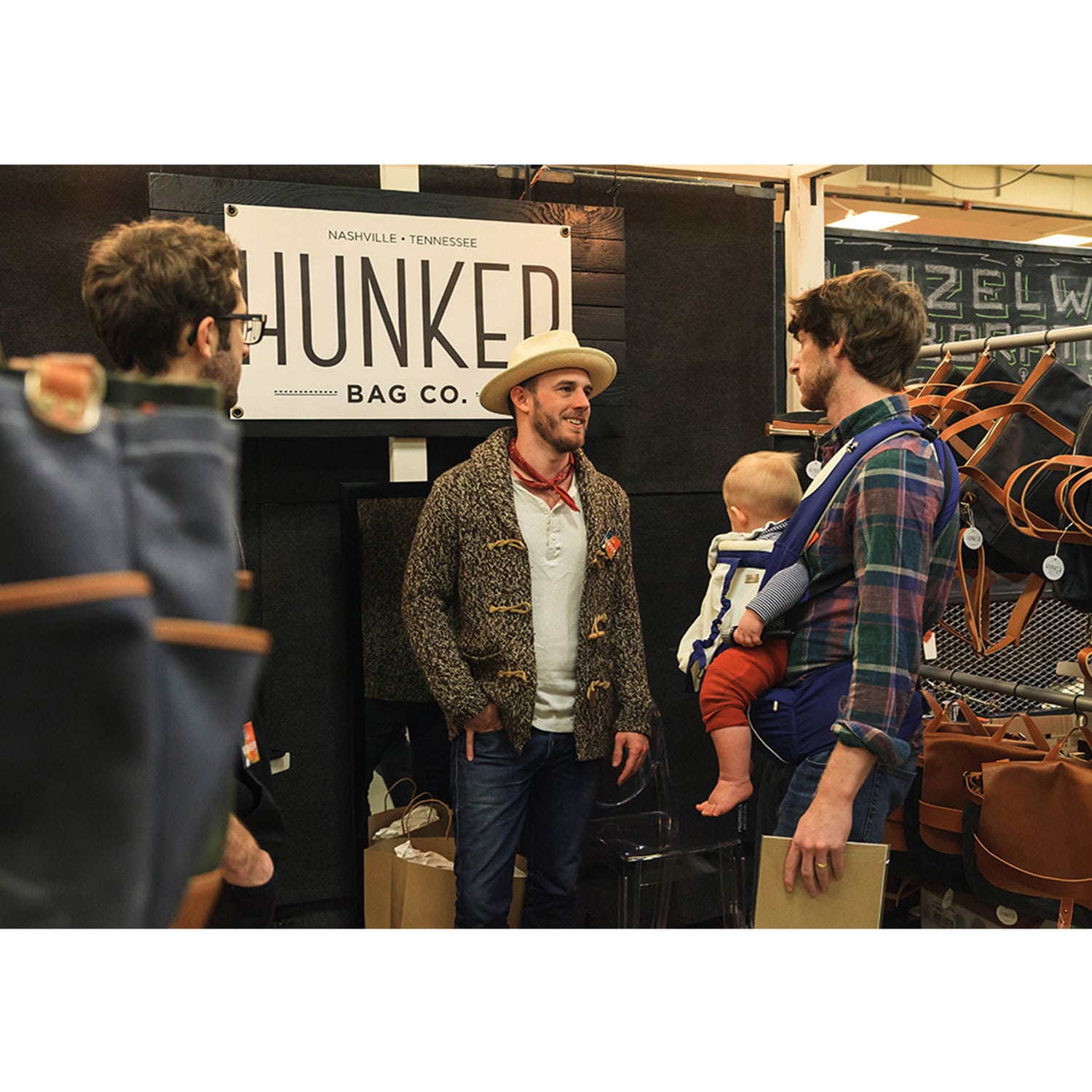 People talking at the Hunker Bag Co. booth, with leather goods and a baby in a carrier