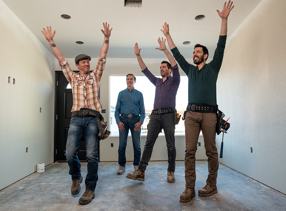 Four men in a partially renovated room, three with tool belts raising their arms in celebration