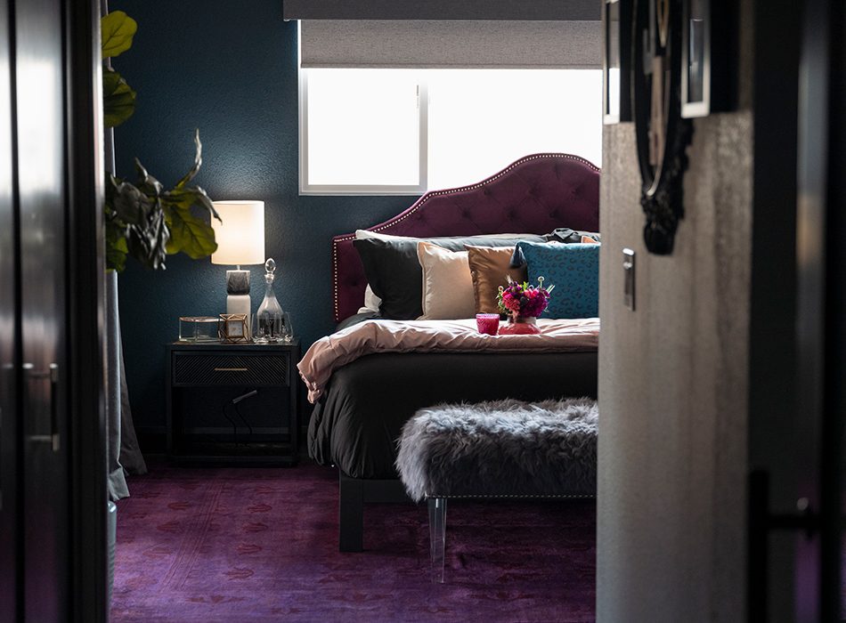 Bedroom with purple headboard, gold and teal pillows, furry bench, and dark purple carpet