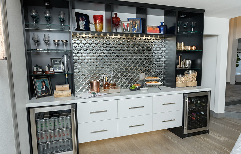 Modern home bar with glass shelves, metallic backsplash, and white drawers